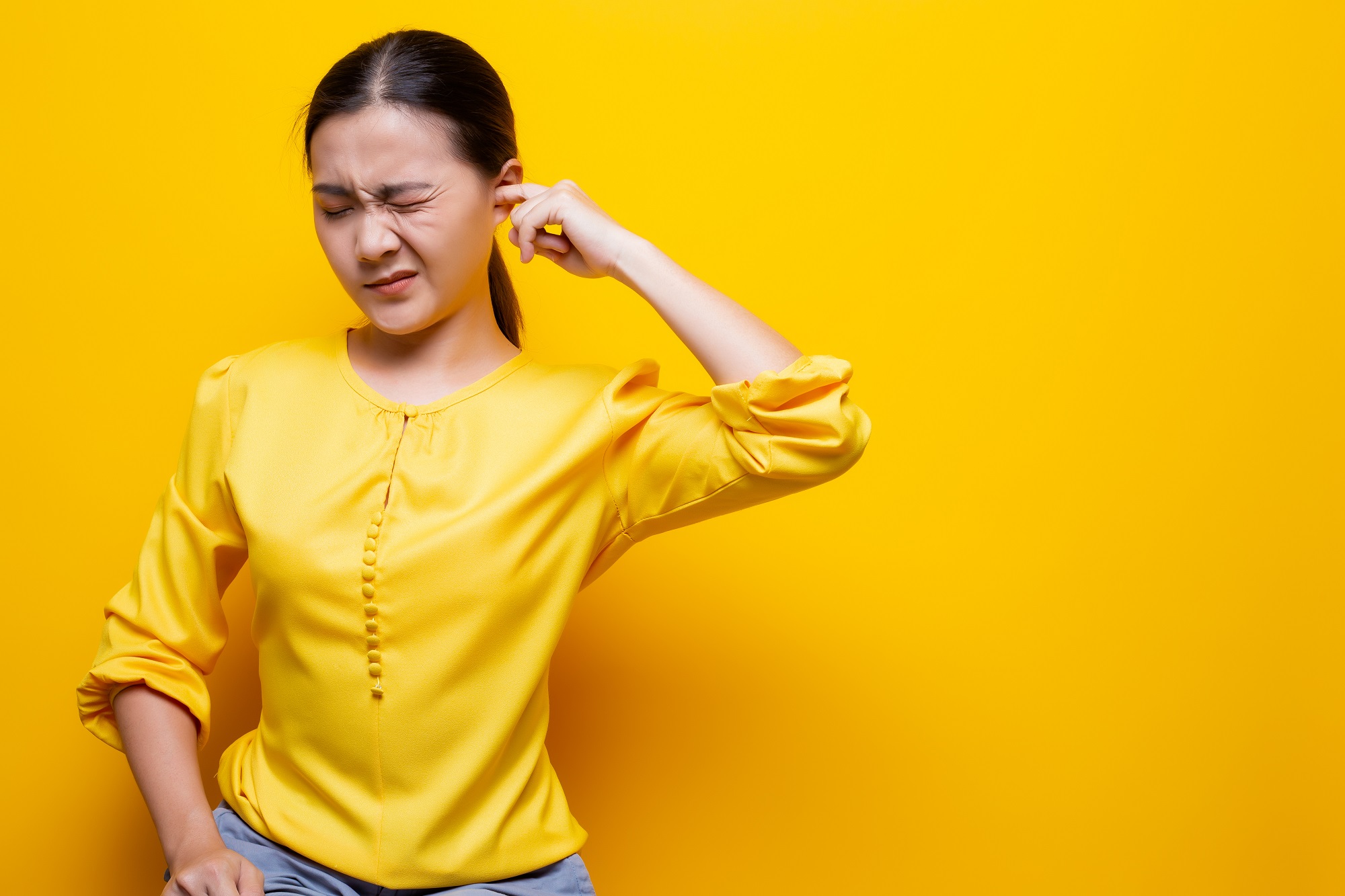 A woman wearing a bright yellow shirt stands in front of a yellow backdrop, putting a finger in her ear for should I go to urgent care for a clogged ear blog.